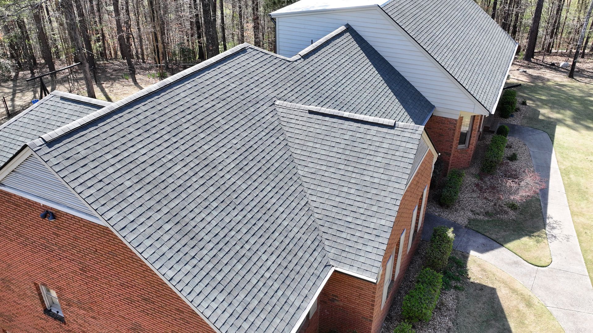 Aerial view of a red brick house with a gray shingled roof, surrounded by trees and a side walkway.