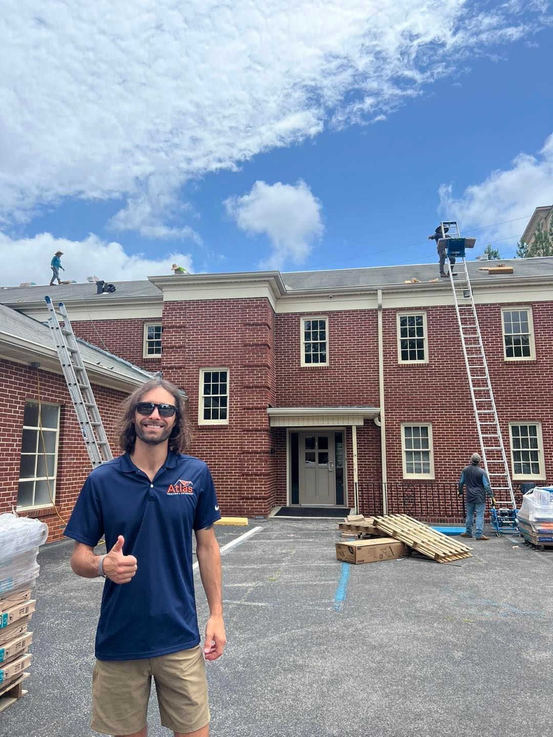 A person wearing a blue polo shirt gives a thumbs up in front of a brick building under repair with workers on the roof.