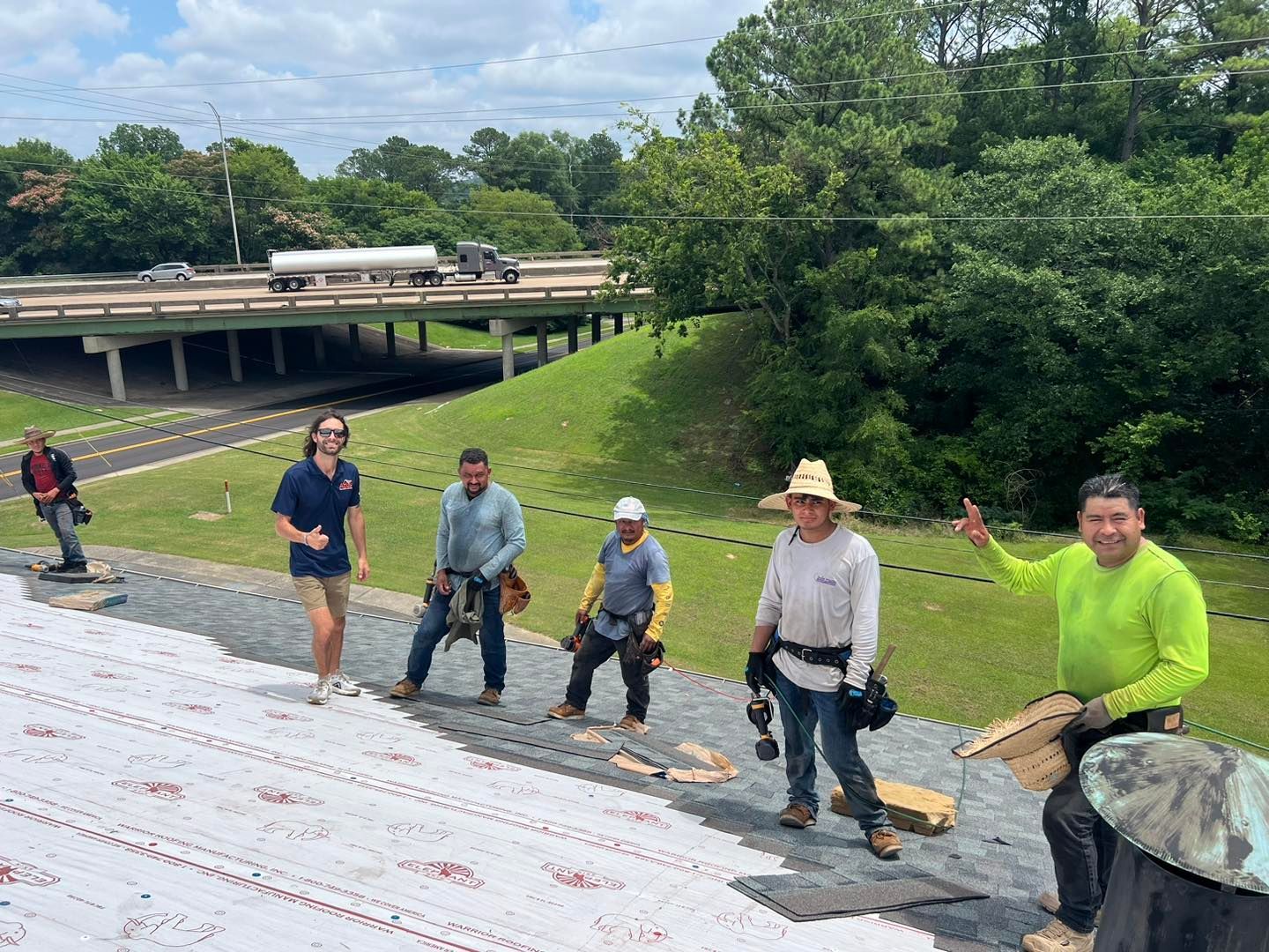 A group of workers on a sloped roof with a highway overpass and trees in the background.