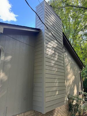 Exterior view of a house side wall featuring a tall, tan-colored wood-paneled chimney chase and a brown roofline.