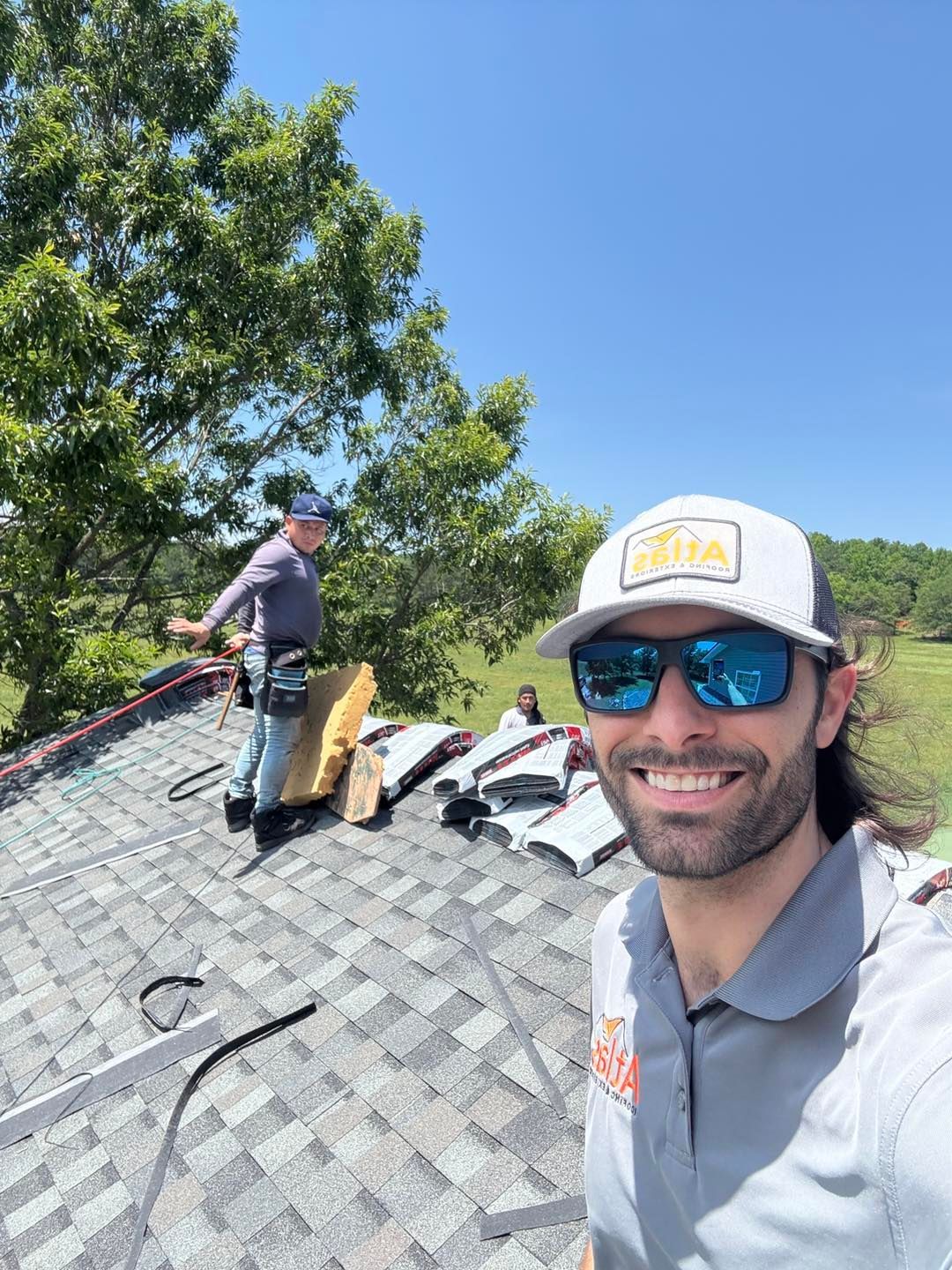 A person wearing a hat and sunglasses smiles in a selfie on a roof, with a coworker nearby working on roofing materials.