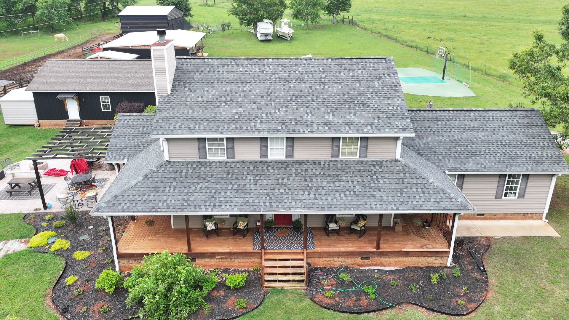 Aerial view of a two-story gray house with a large front porch, a separate dark building, and a backyard with a court.