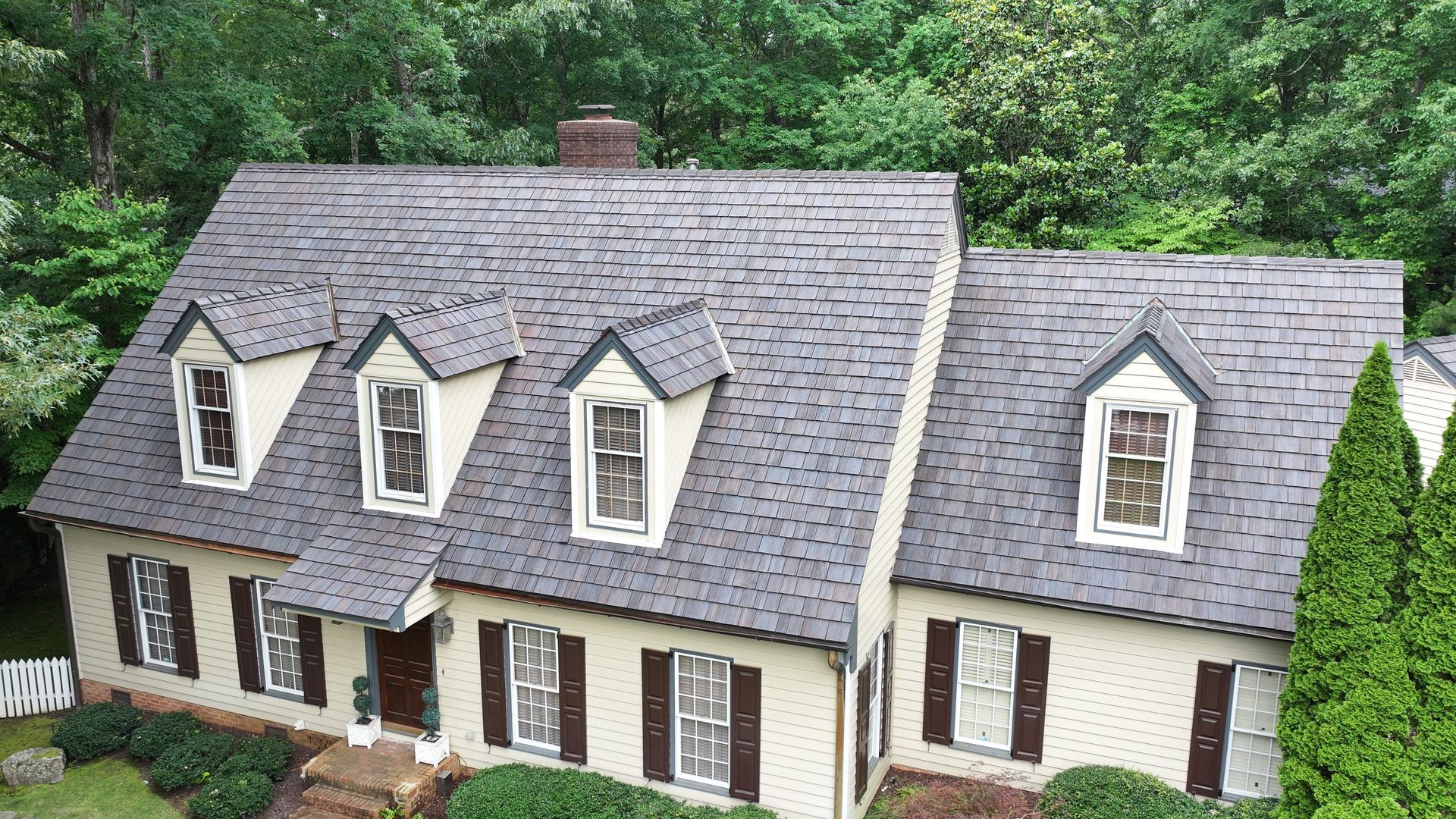 A light-colored two-story home with a dark shingled roof, four dormer windows, and dark shutters, surrounded by trees.