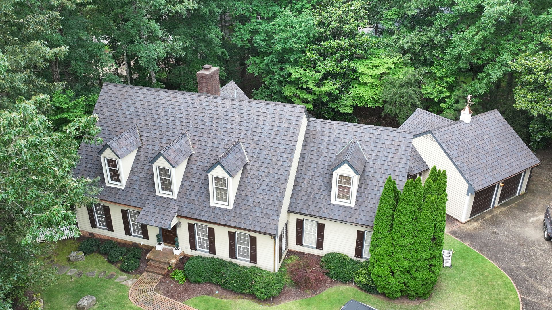 A cream-colored, two-story house with a grey shingled roof, three dormers, and an attached garage surrounded by trees.