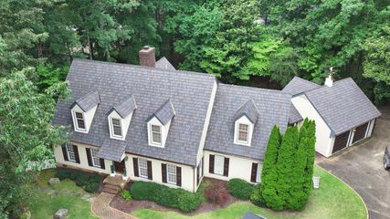 An aerial view of a tan two-story house with a gray shingle roof, dormer windows, shutters, and a detached garage.