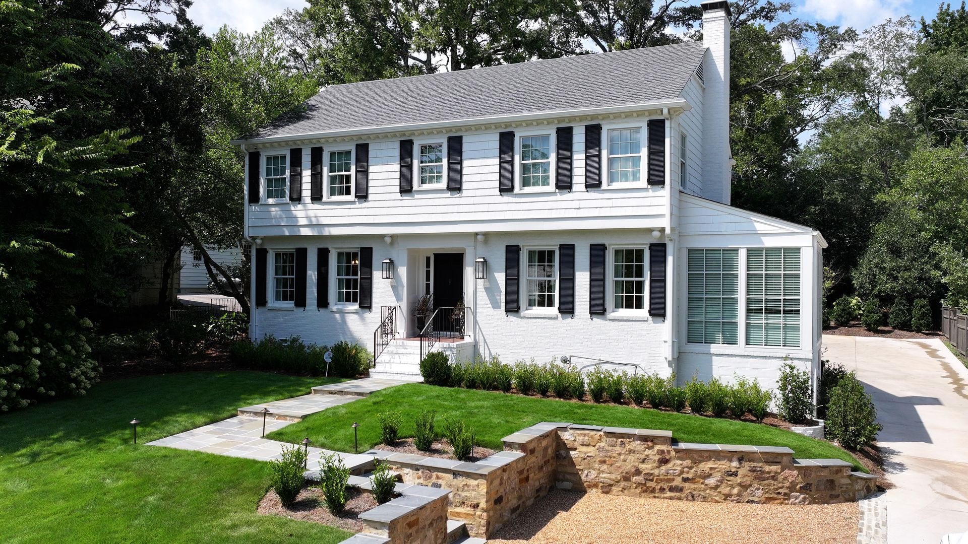 A two-story white colonial-style house with black shutters, a stone retaining wall, and a green lawn in a wooded setting.