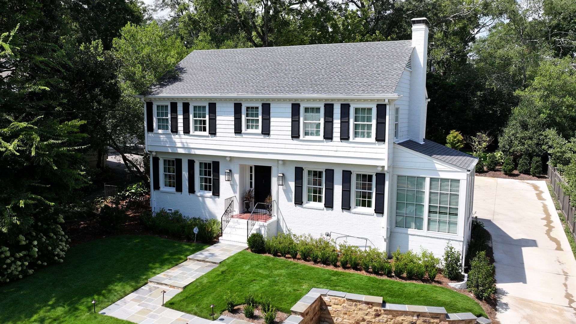 A two-story white colonial-style house with black shutters, a shingled roof, a front lawn, and a paved driveway.