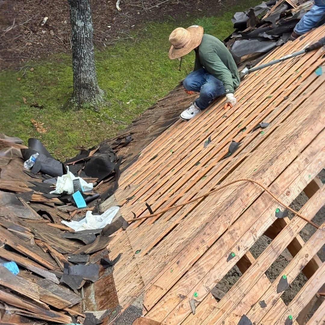 A person wearing a wide-brimmed hat works on a wooden roof structure, removing old shingles and debris.