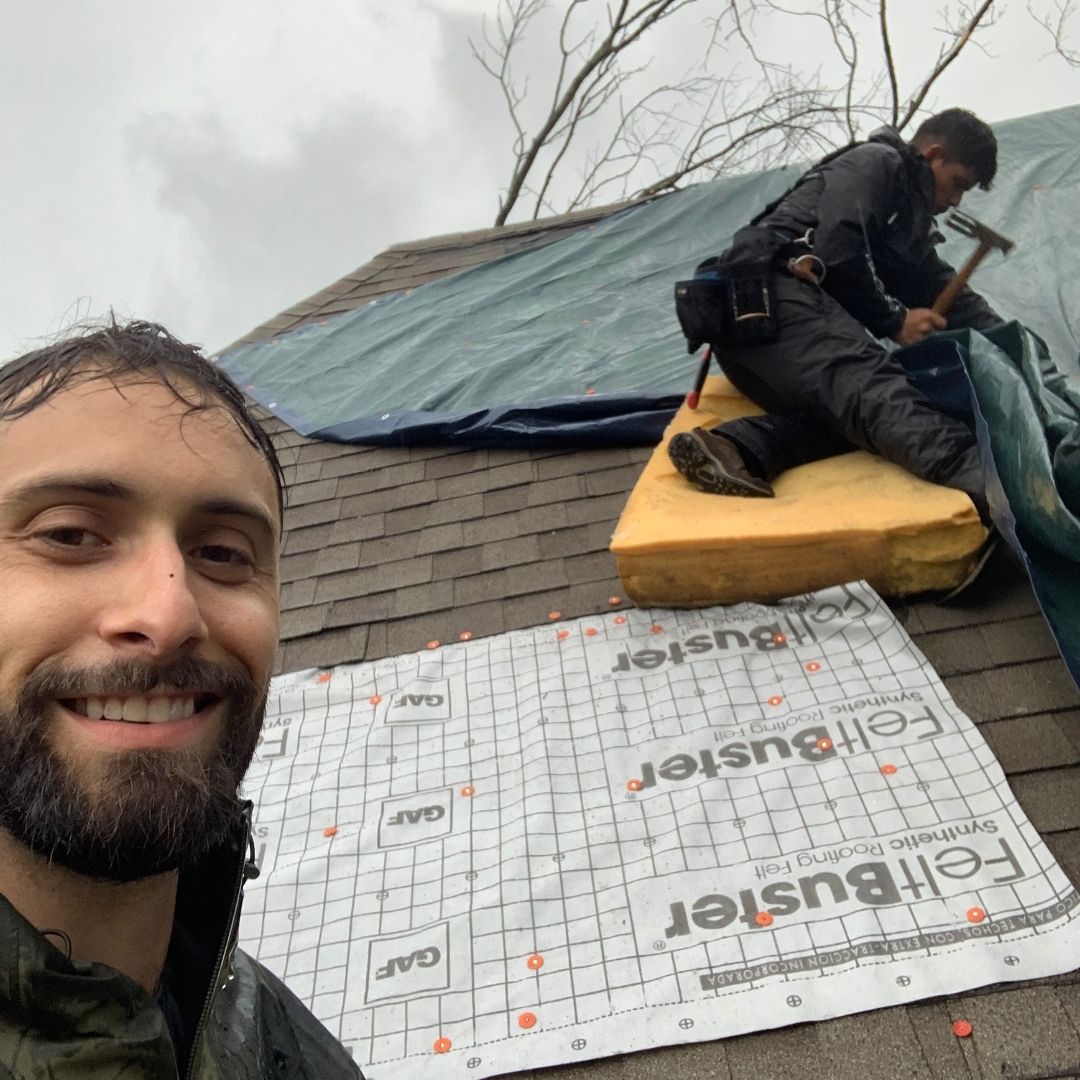A smiling man in the foreground and a coworker using a hammer on a roof covered in tarp and FeltBuster underlayment.