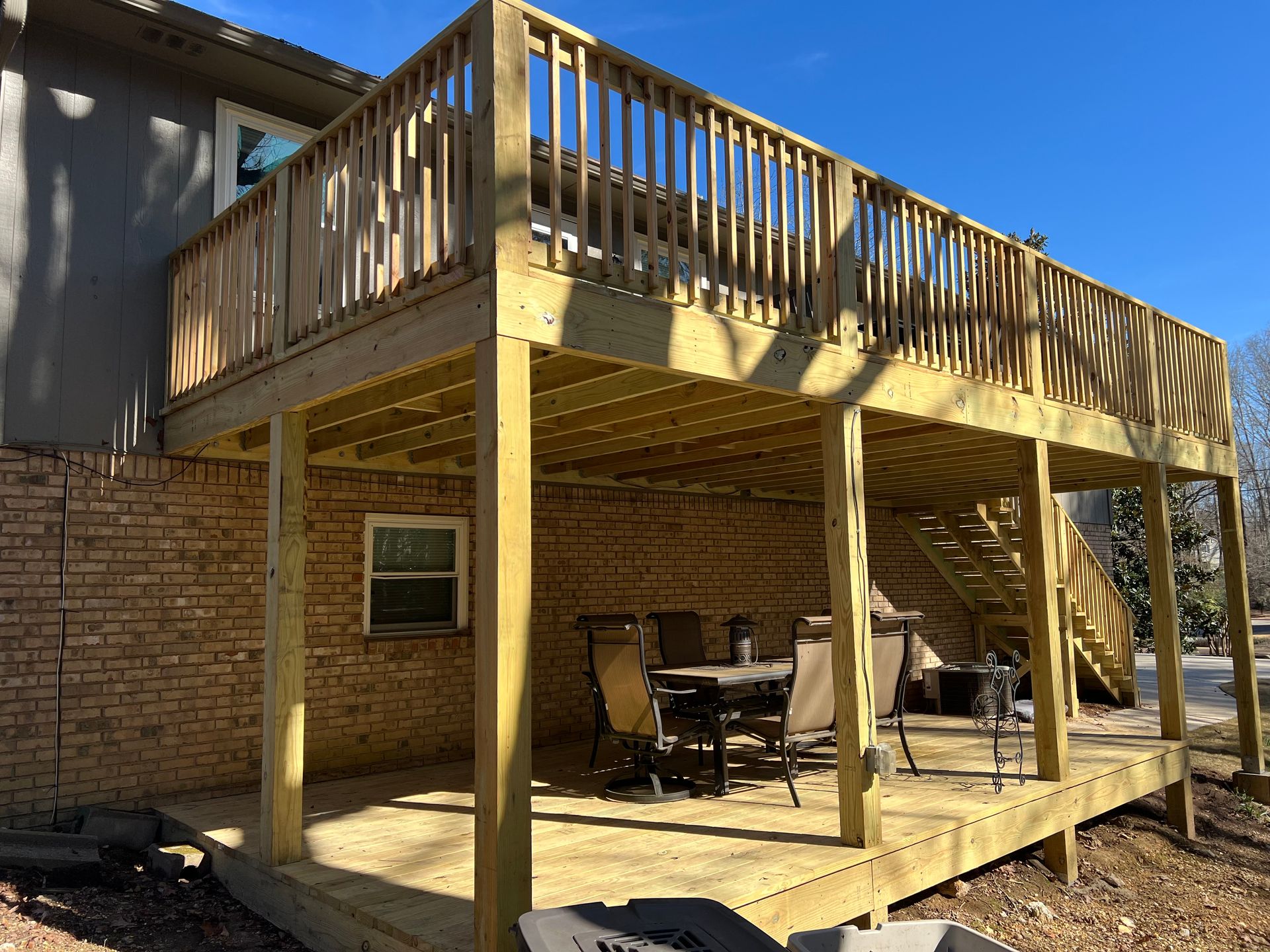 A wooden deck and patio area attached to the back of a brick house, featuring outdoor seating under the raised deck.