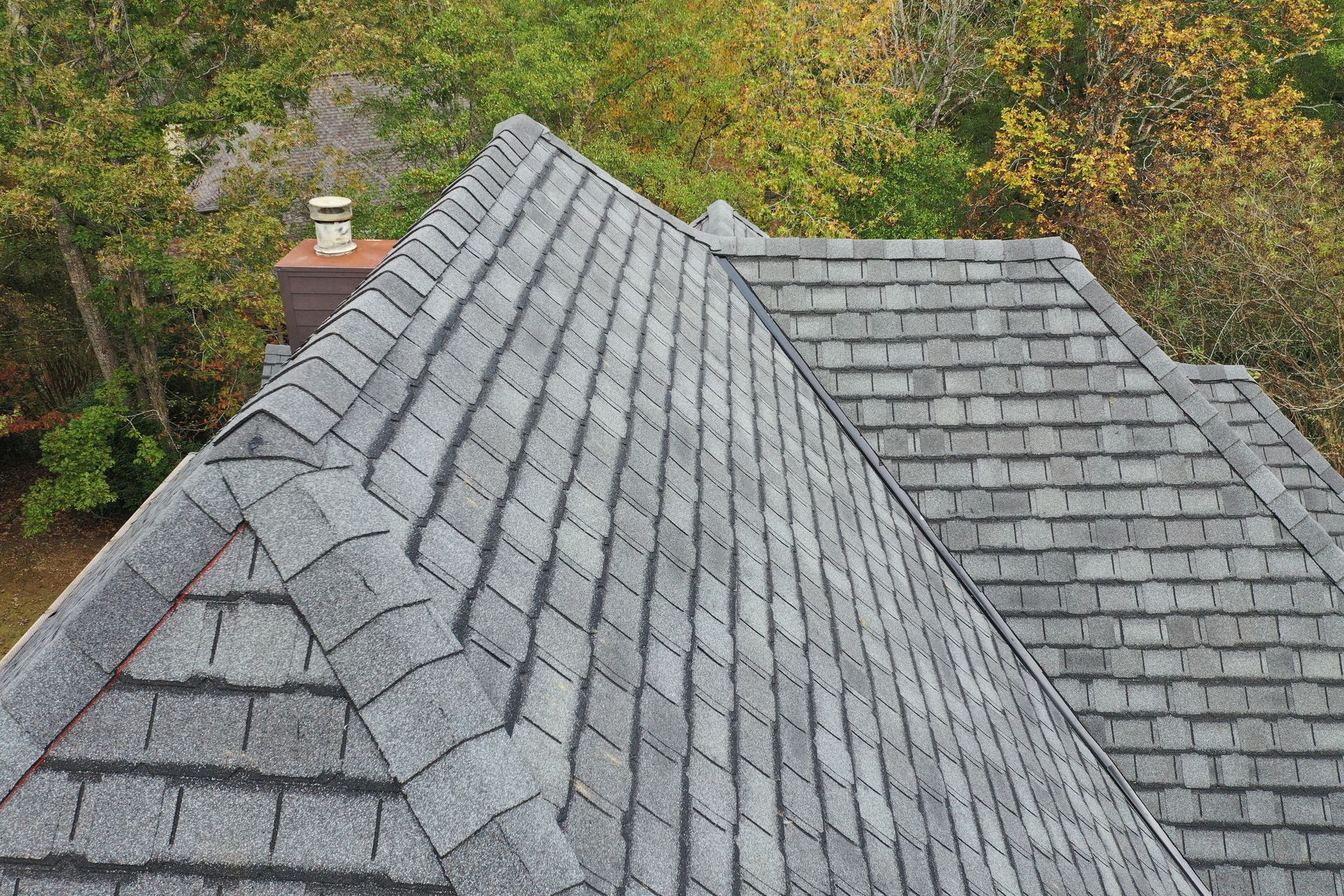 High-angle view of a residential roof with grey asphalt shingles, a brick chimney, and surrounding green and yellow trees.