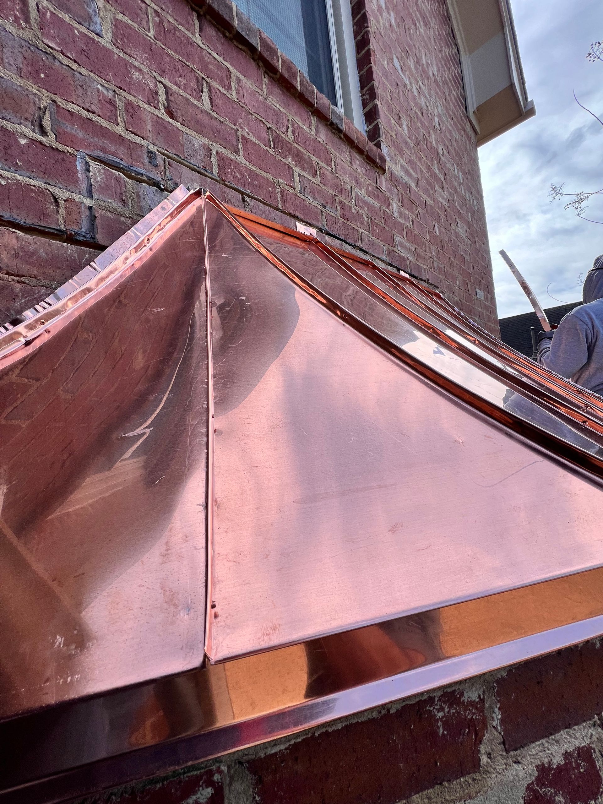 A close-up of a new, shiny copper roof cap installed against a brick wall exterior.