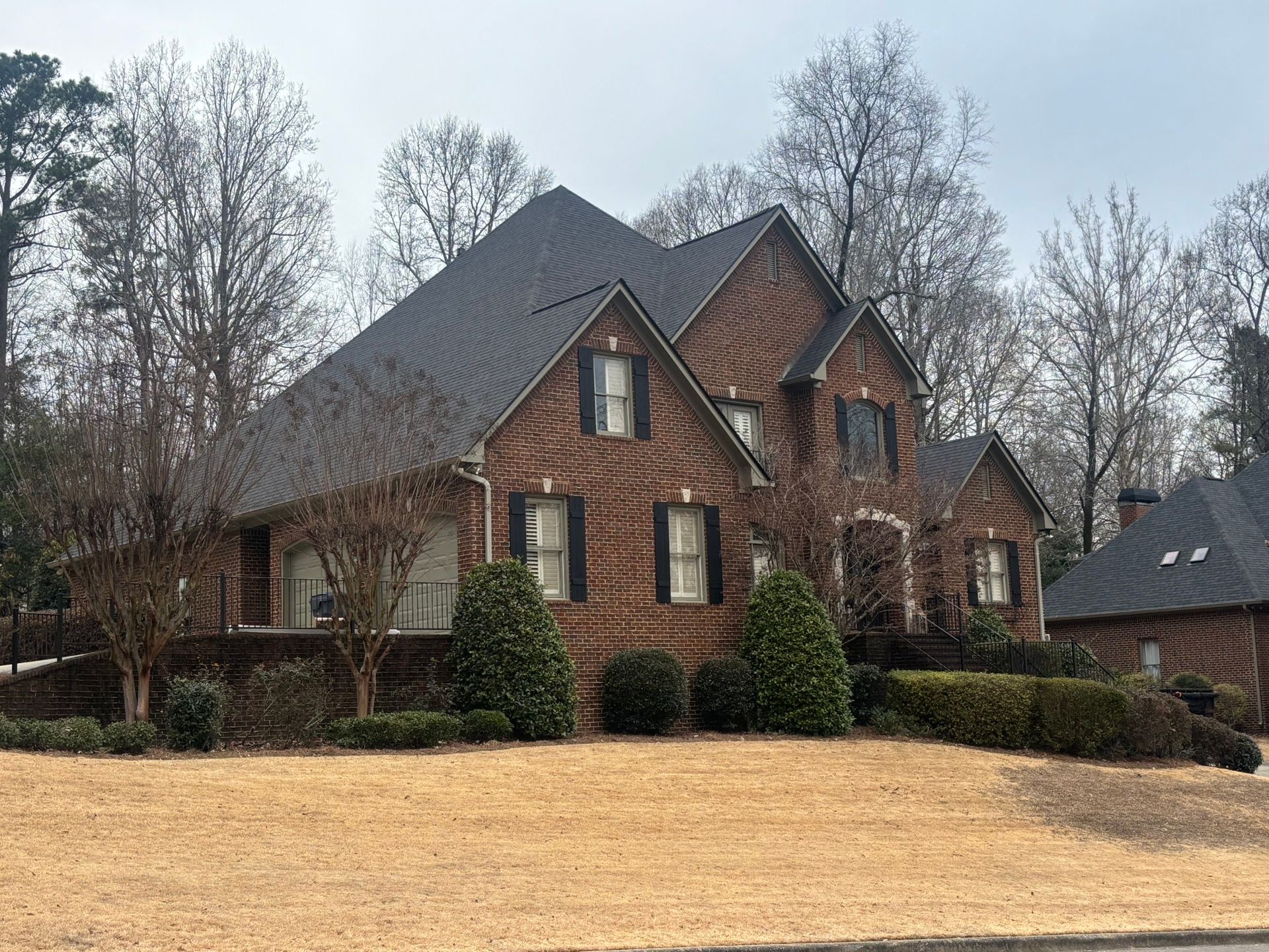 A two-story brick house with a dark gabled roof and black shutters, situated on a grassy hill with trees in the background.