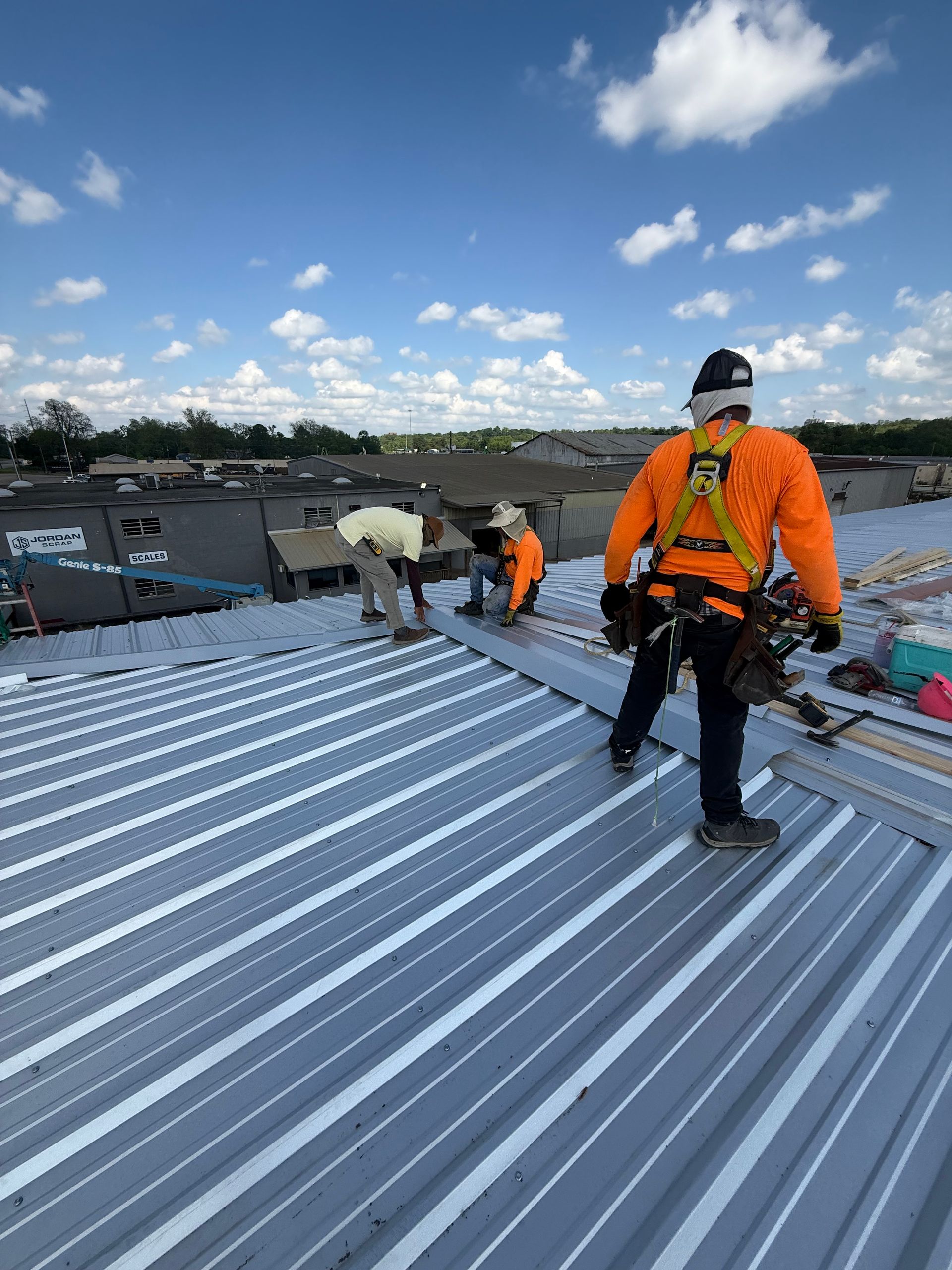 Three construction workers in safety gear install light-colored metal roofing panels under a bright, sunny sky.