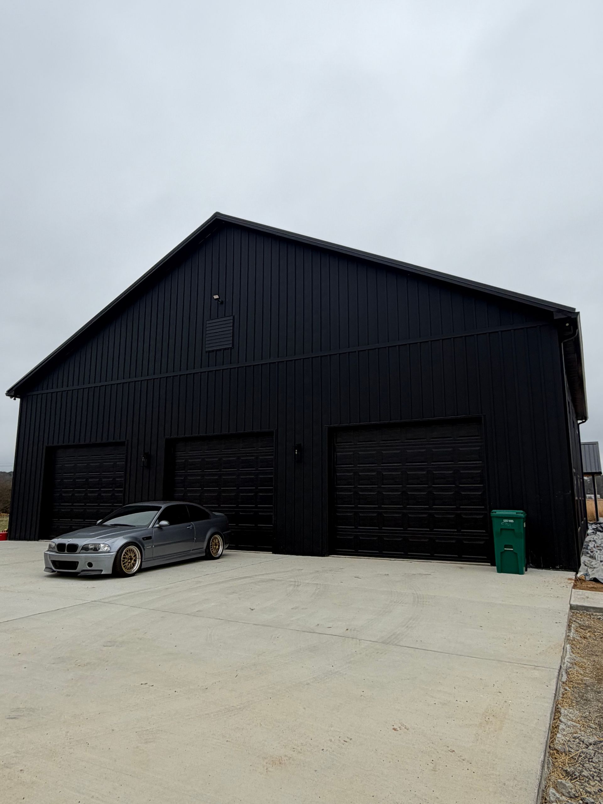 A silver BMW sedan parked in front of a modern, black metal-sided garage building with three bay doors under a gray sky.