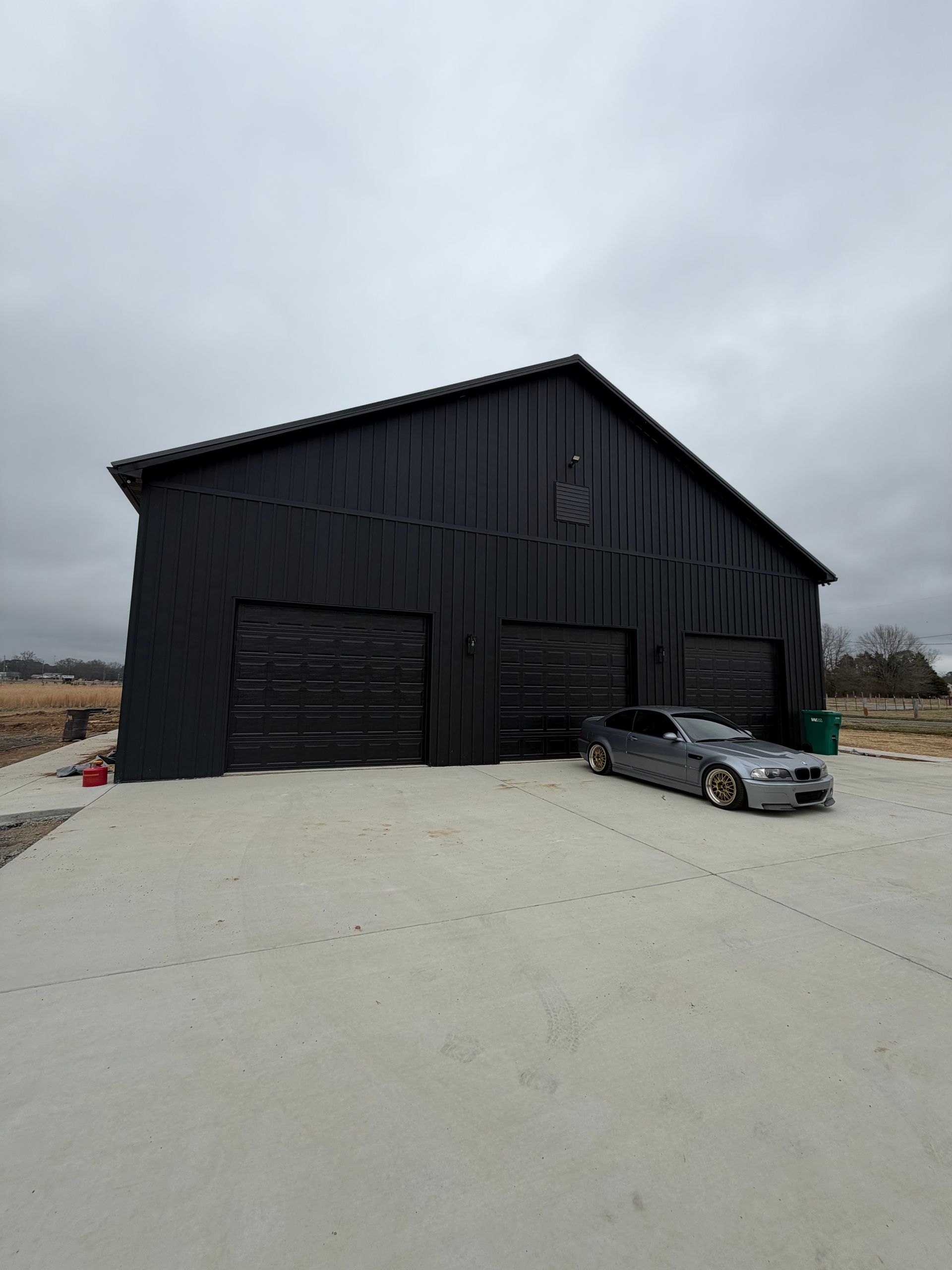 A silver car parked on a concrete driveway in front of a large, black, three-bay metal garage under a cloudy sky.