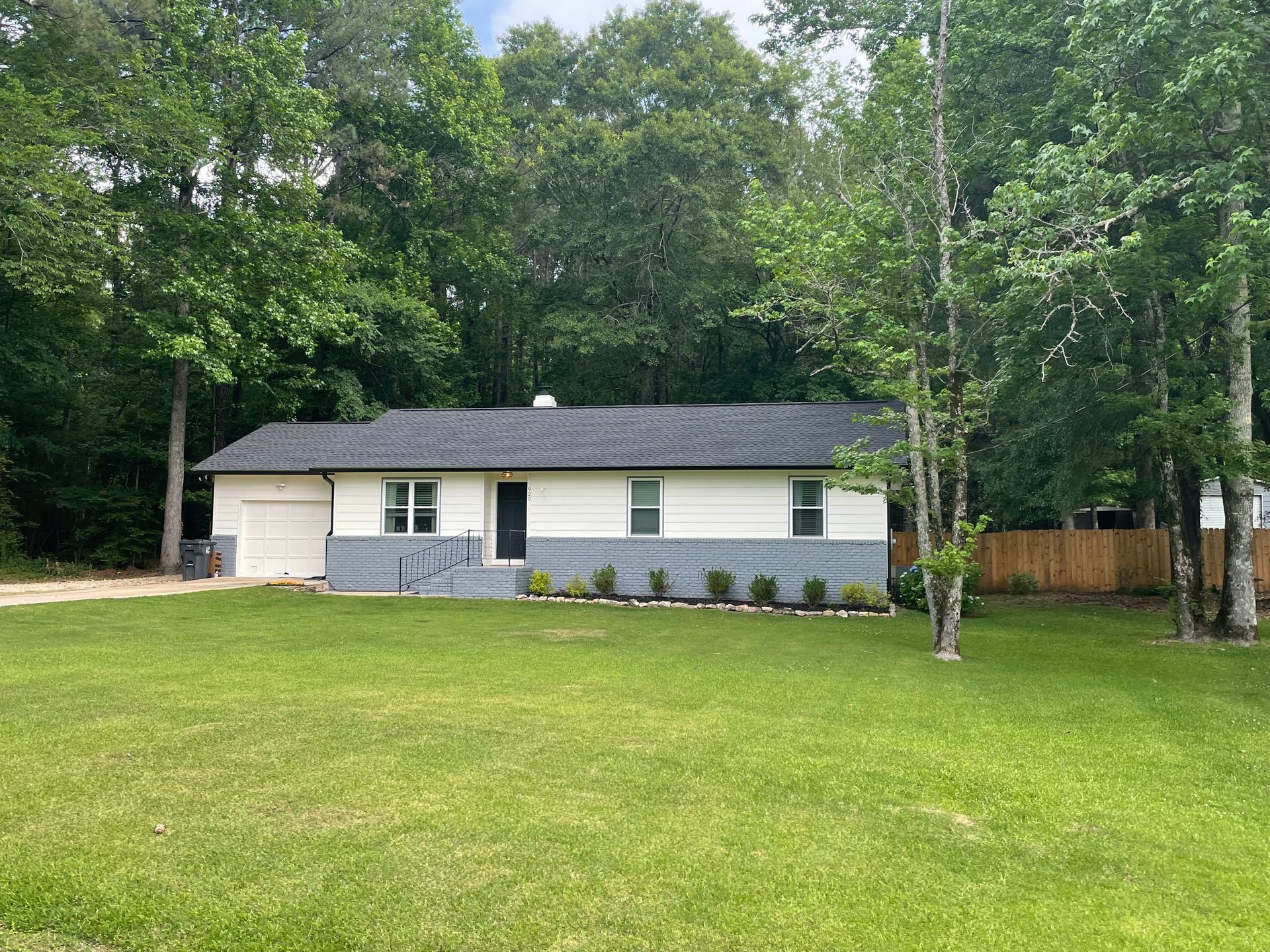 A one-story house with white siding and gray stone base, black roof, and front door, set in a yard with trees.