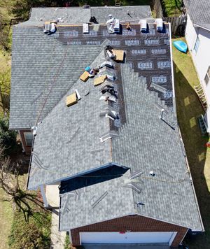 A high-angle view of a house roof undergoing repairs, showing sections of new dark gray shingles and exposed black felt.