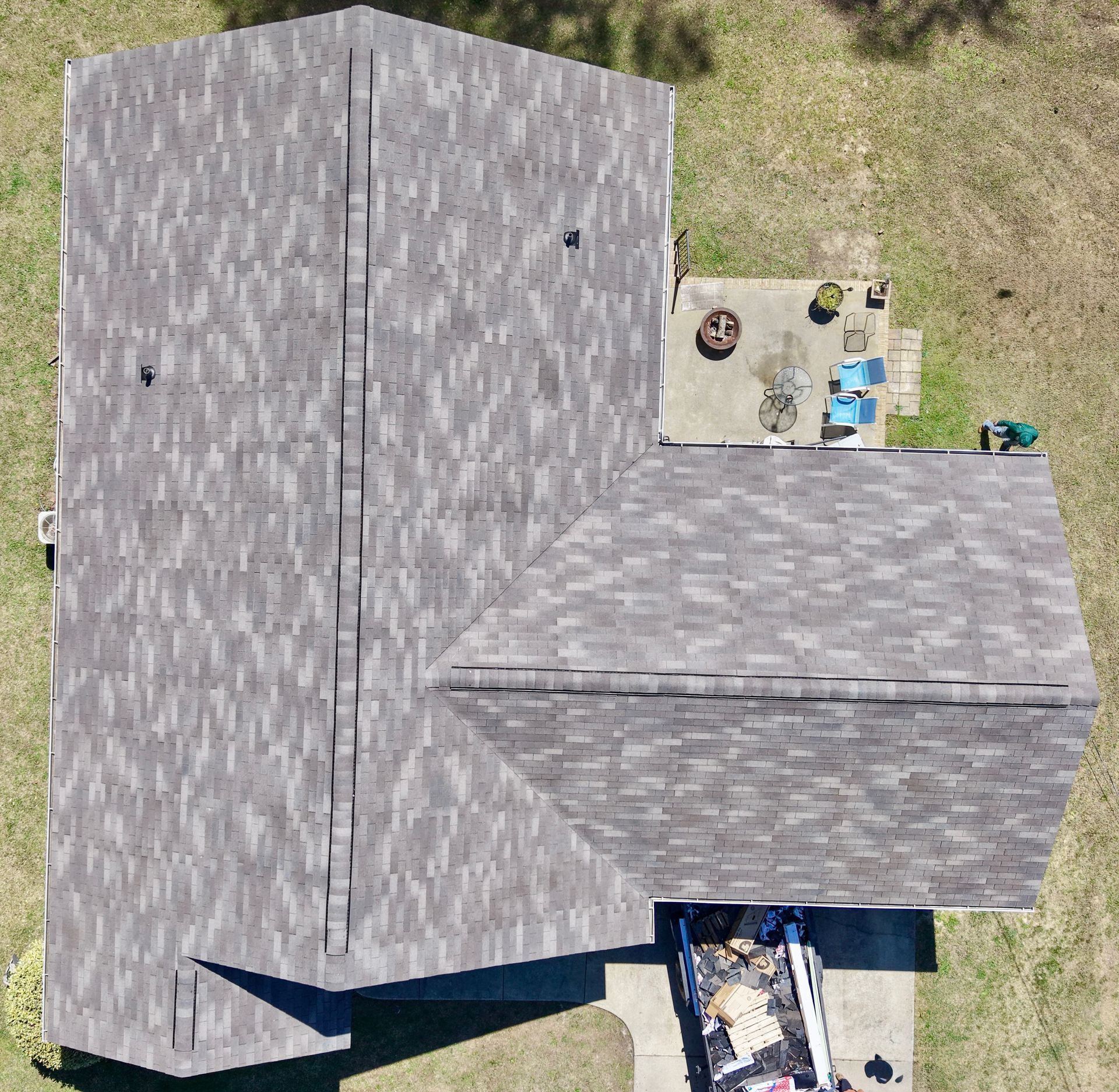 A top-down aerial view of a dark grey shingled roof on a house, with a patio and a construction dumpster visible below.