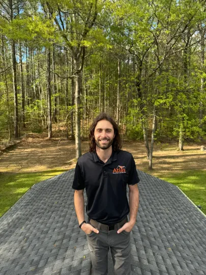 A person with long hair stands on a dark shingled roof outdoors, smiling with hands in pockets, backed by tall trees.