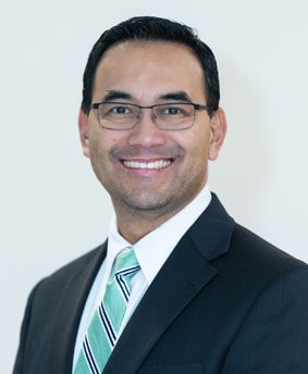 Smiling man in a dark suit, glasses, and striped tie against a light background
