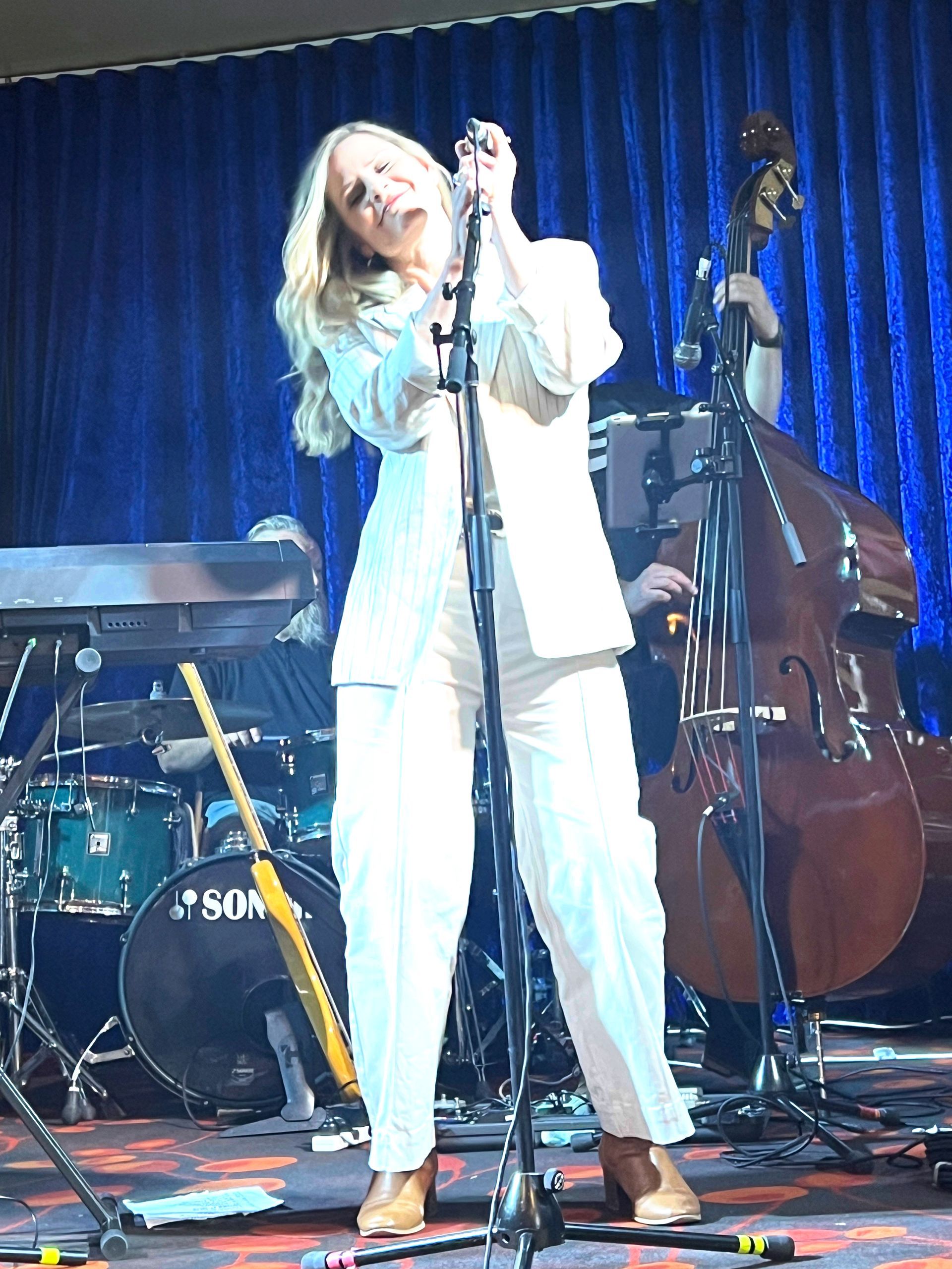 Blonde singer in white suit, performing on stage with band, blue curtain backdrop.