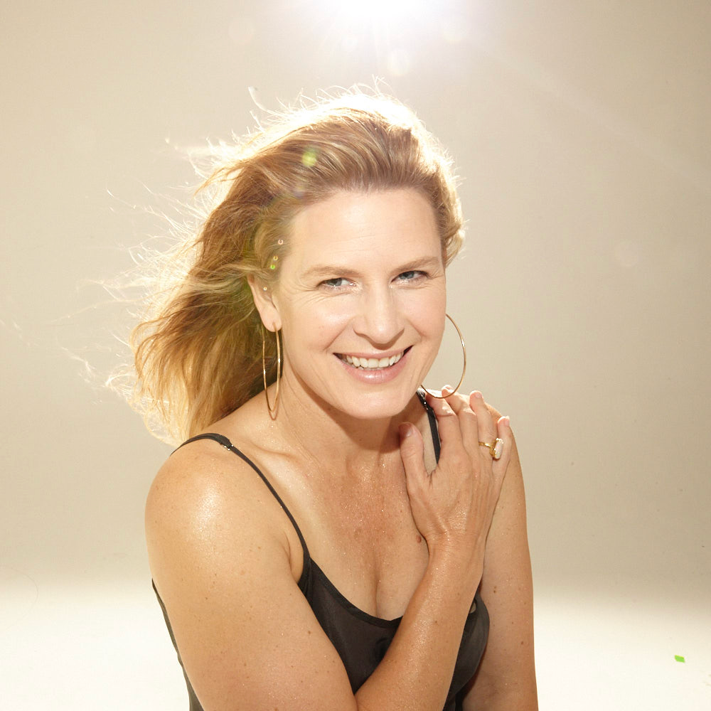 Woman smiling, arm on shoulder, wearing a black top, large hoop earrings, and a ring, with light background.