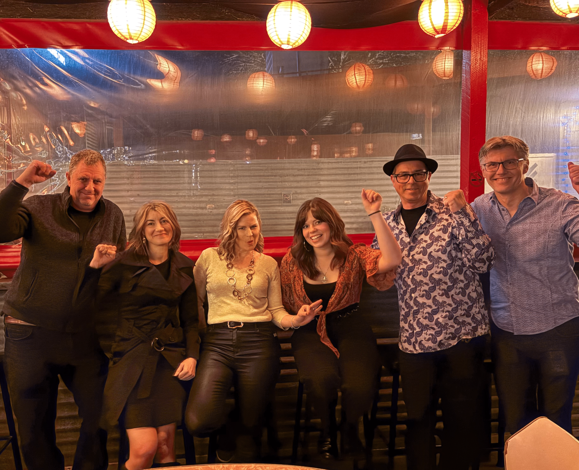 Six people at an outdoor restaurant, clenching fists in celebration. Lights hang overhead, red canopy.