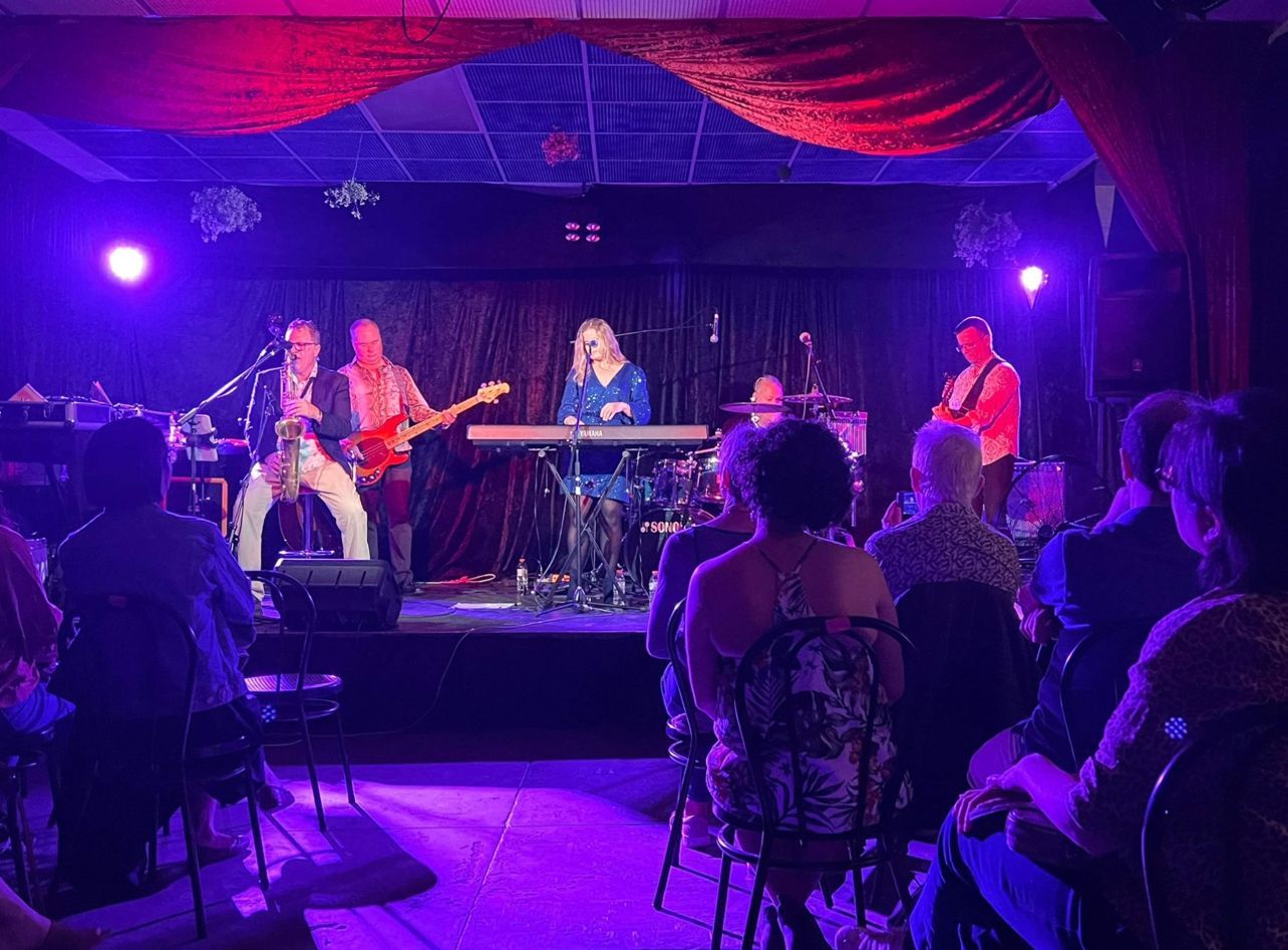 Band performing on stage in a dimly lit venue, audience watching.