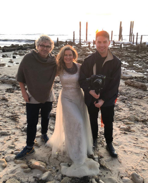 Woman in wedding dress with two people on a beach at sunset, camera in hand.
