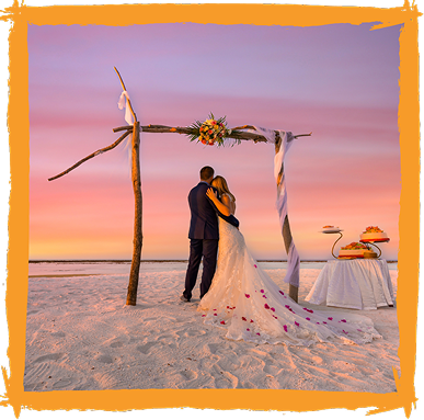 Bride and groom embrace beneath a floral arch on a beach at sunset.