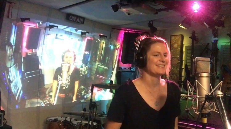 Woman singing into a microphone in a studio with a projected image on a wall. She is smiling.