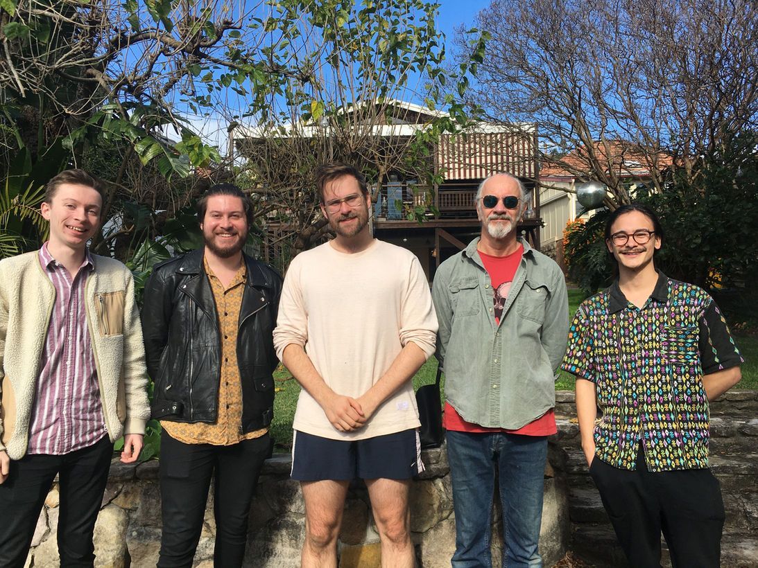 Five people standing outside, smiling. The man in the middle wearing shorts. Trees and a house in the background.