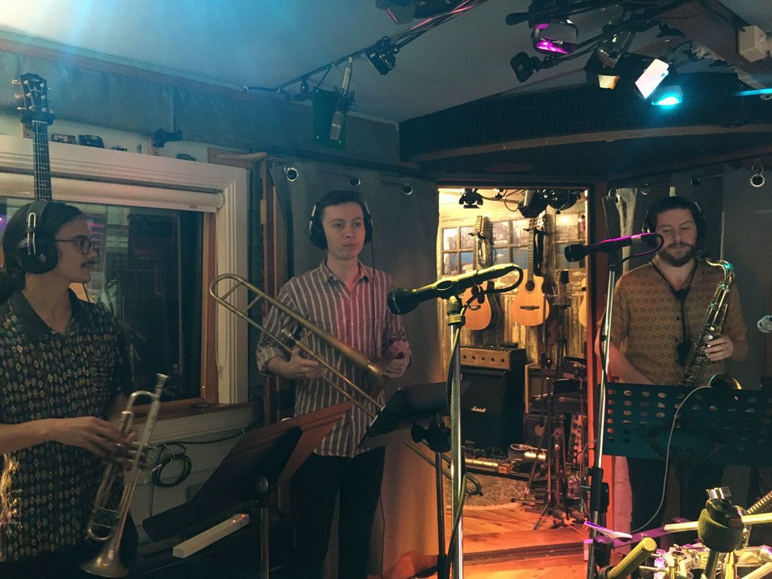 Three musicians in a recording studio. A trumpet, trombone, and saxophone player stand by mics, ready to play.