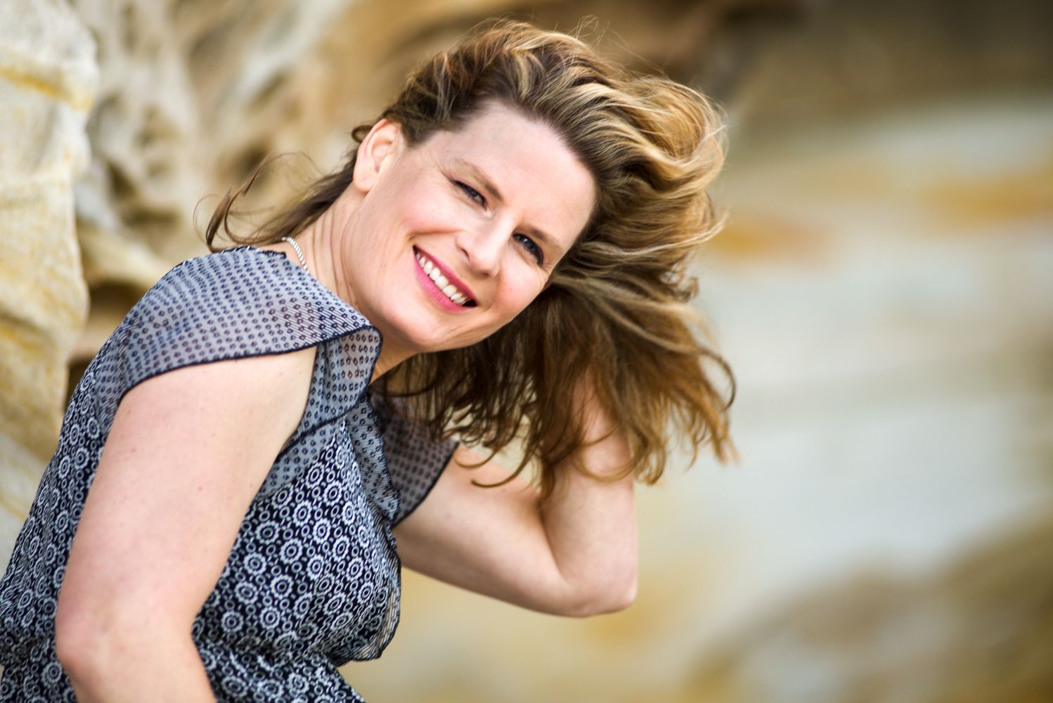 Woman smiles, adjusts hair, outdoors. She wears a patterned top with a rocky background.