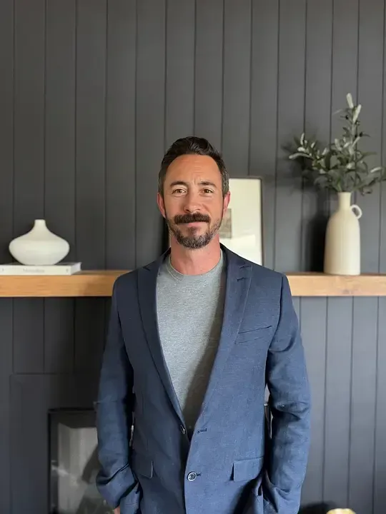 Man in blue suit and gray shirt, standing in front of a shelf with decor, dark gray wall.