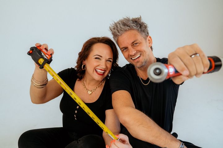 Couple holding a tape measure and saw, smiling with arms outstretched against a white background.