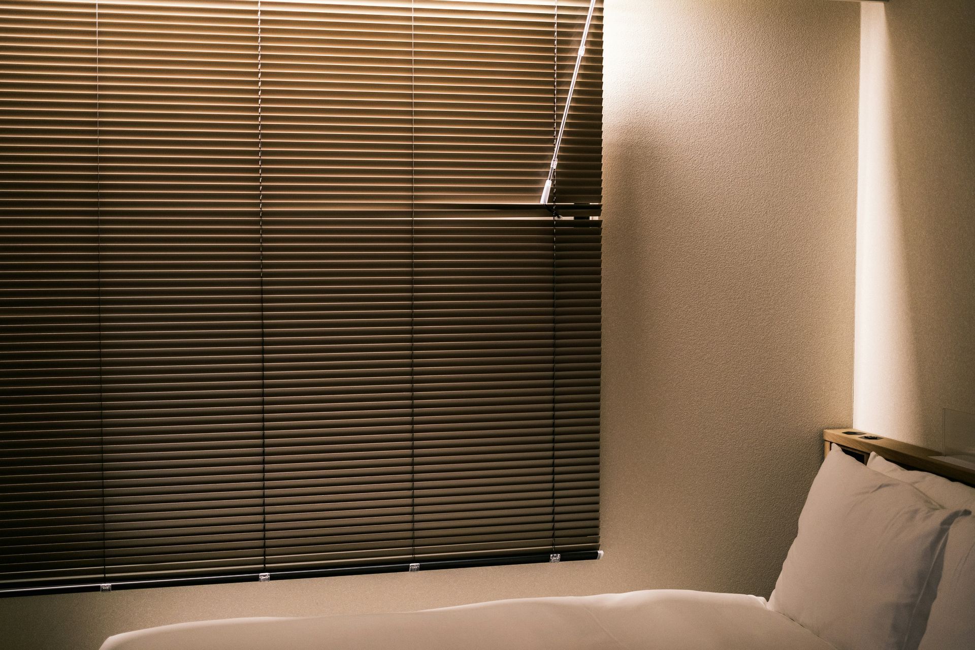 Window with closed blinds casting striped shadows over a bed with white pillows.