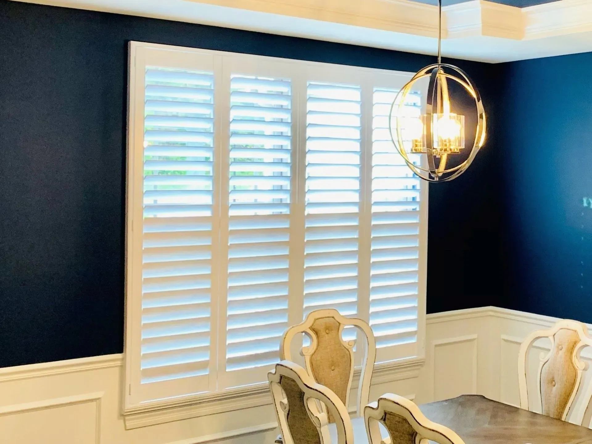 A dining room with a table and chairs and a window with white shutters.