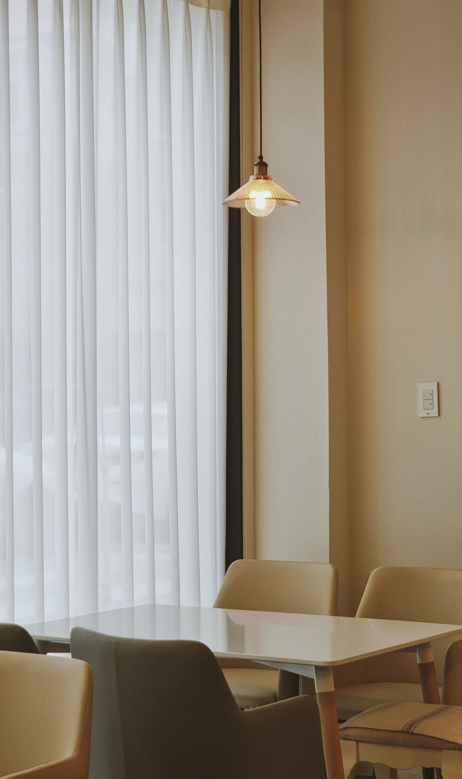 Dining area with white curtains, pendant light, and table with chairs. Neutral tones.