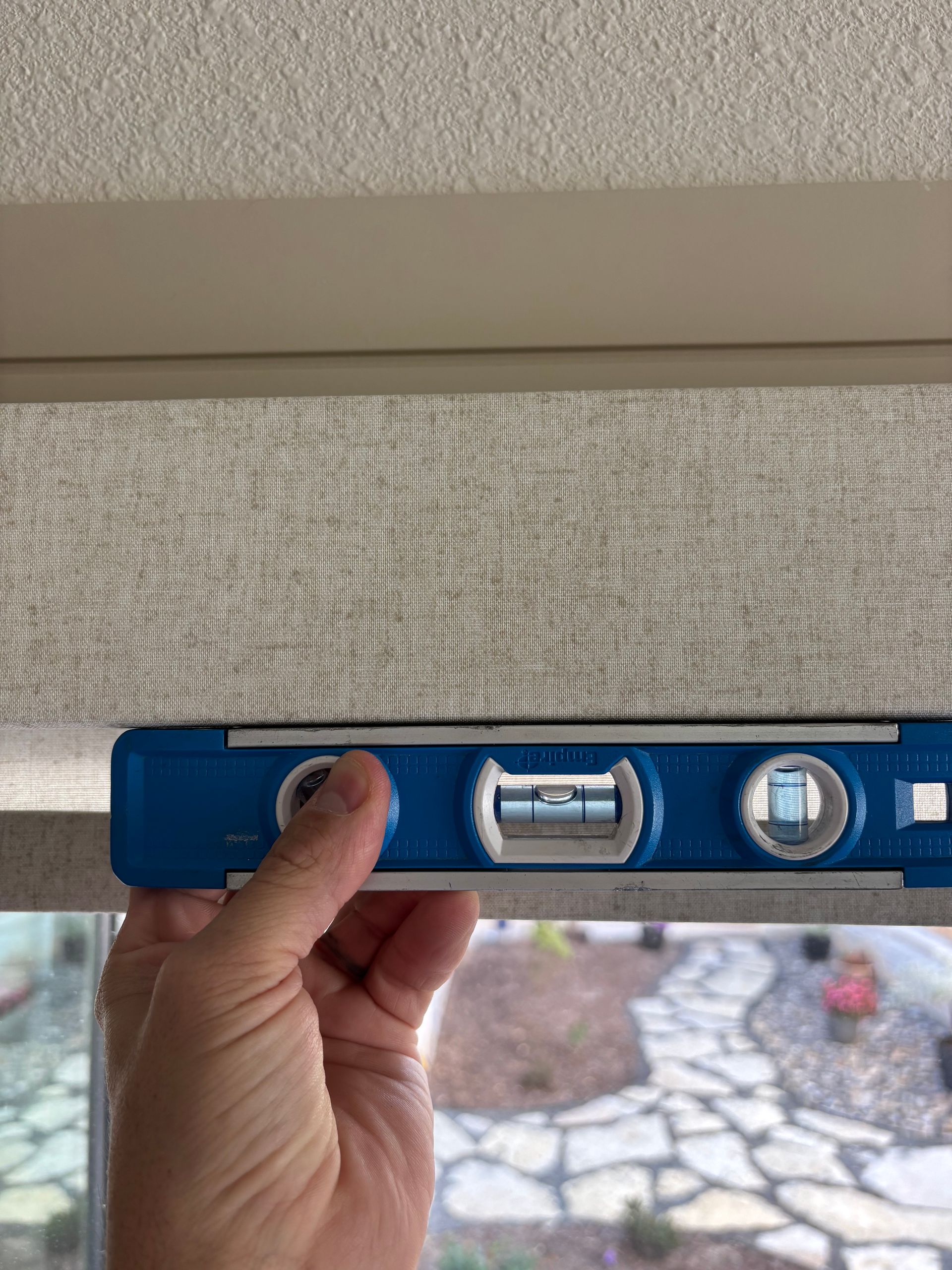Hand holding a blue level to check a horizontal surface of a blind in front of a window.