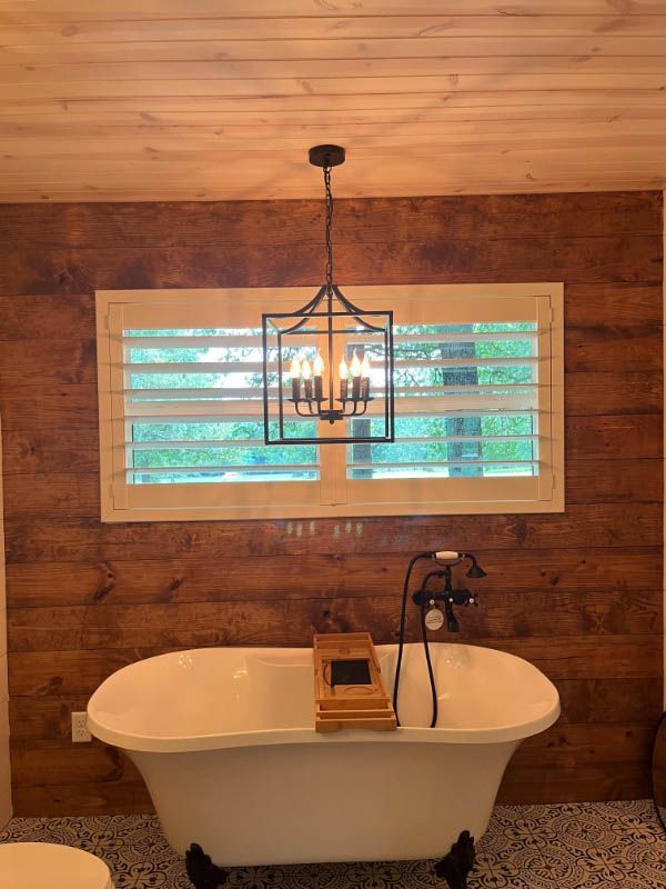 A rectangular window with shutters is in an elegant bathroom over a claw foot tub. 