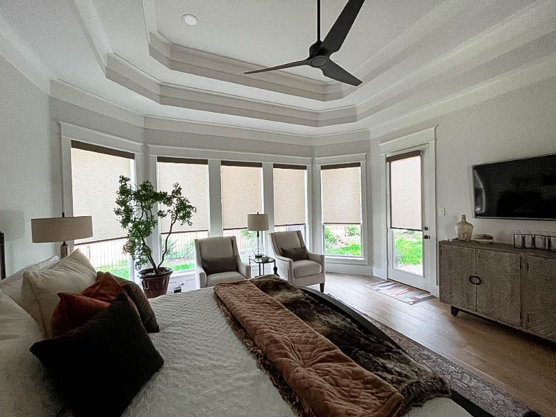 A large bowed window with roller shades is in a large, white bedroom. 