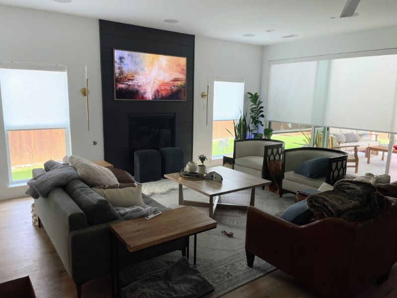 A living room with a wall-mounted television has a wall of windows with roller shades. 