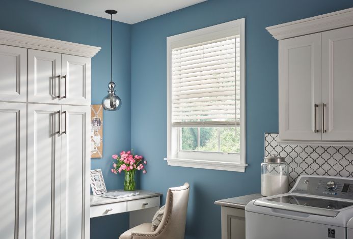 A blue kitchen with white trim has a window with white wood blinds. 