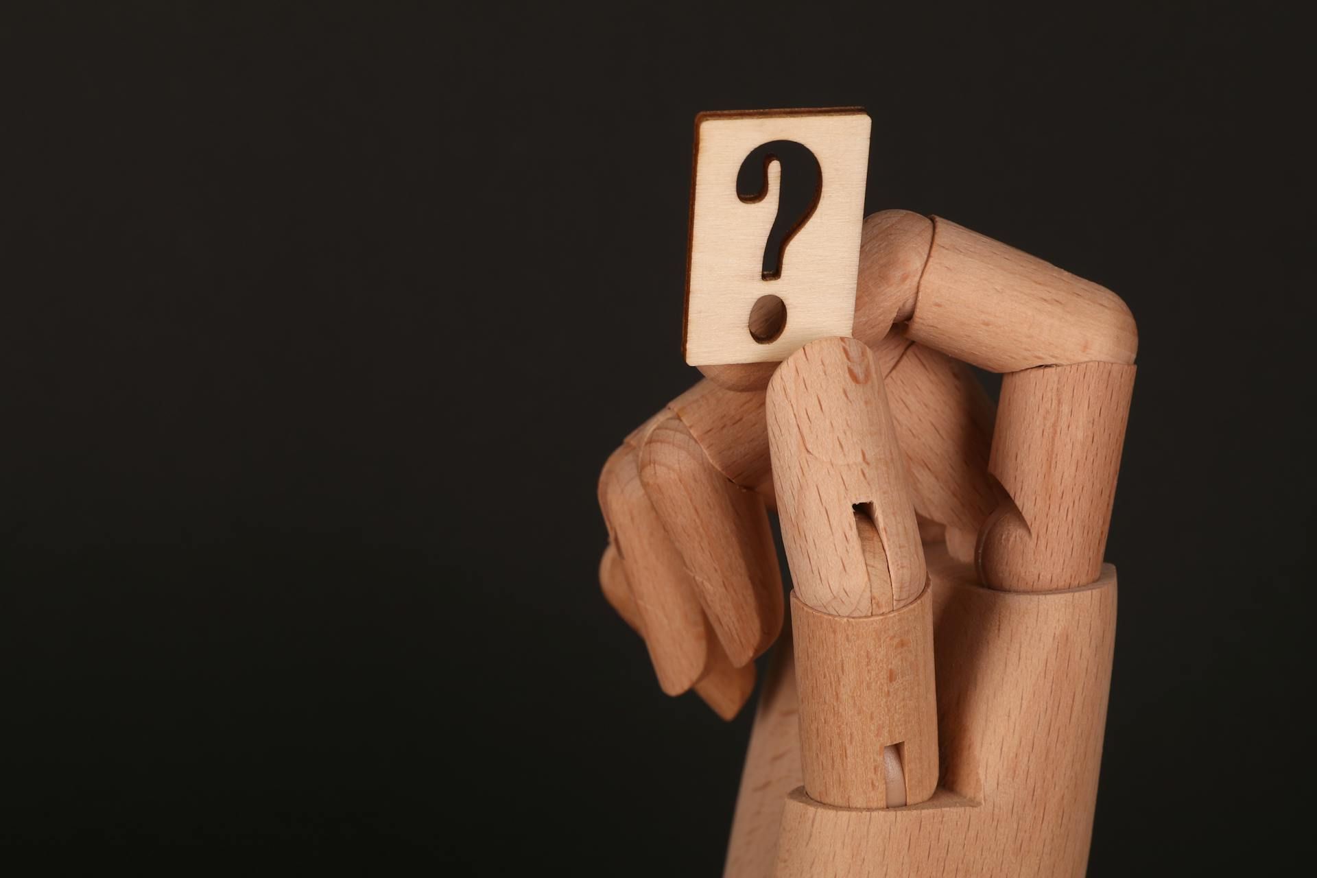 A wooden mannequin hand holds a small card with a question mark on it against a black background.