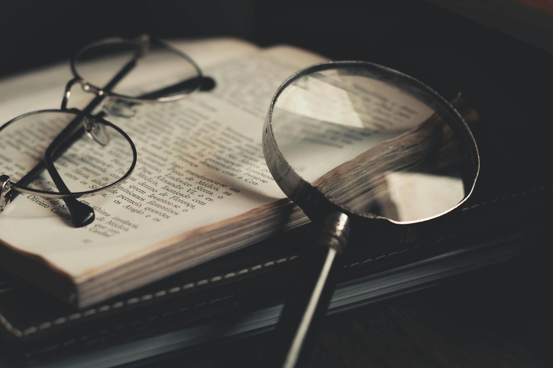 A magnifying glass resting on an open vintage book, with a pair of round wire-rimmed glasses placed on the pages.