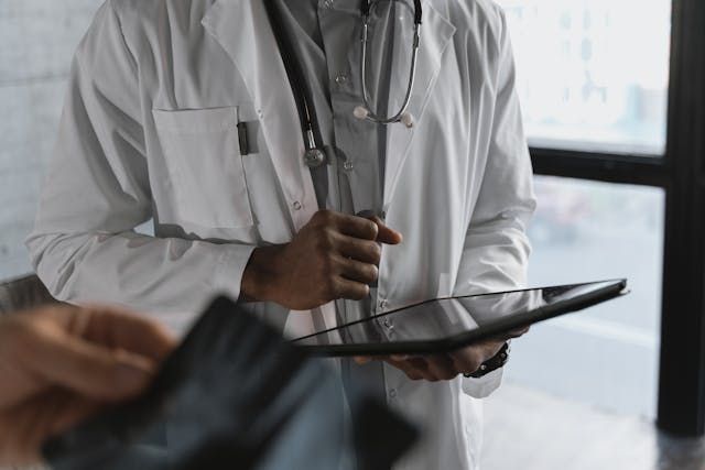A person in a white lab coat with a stethoscope around their neck holds a digital tablet.