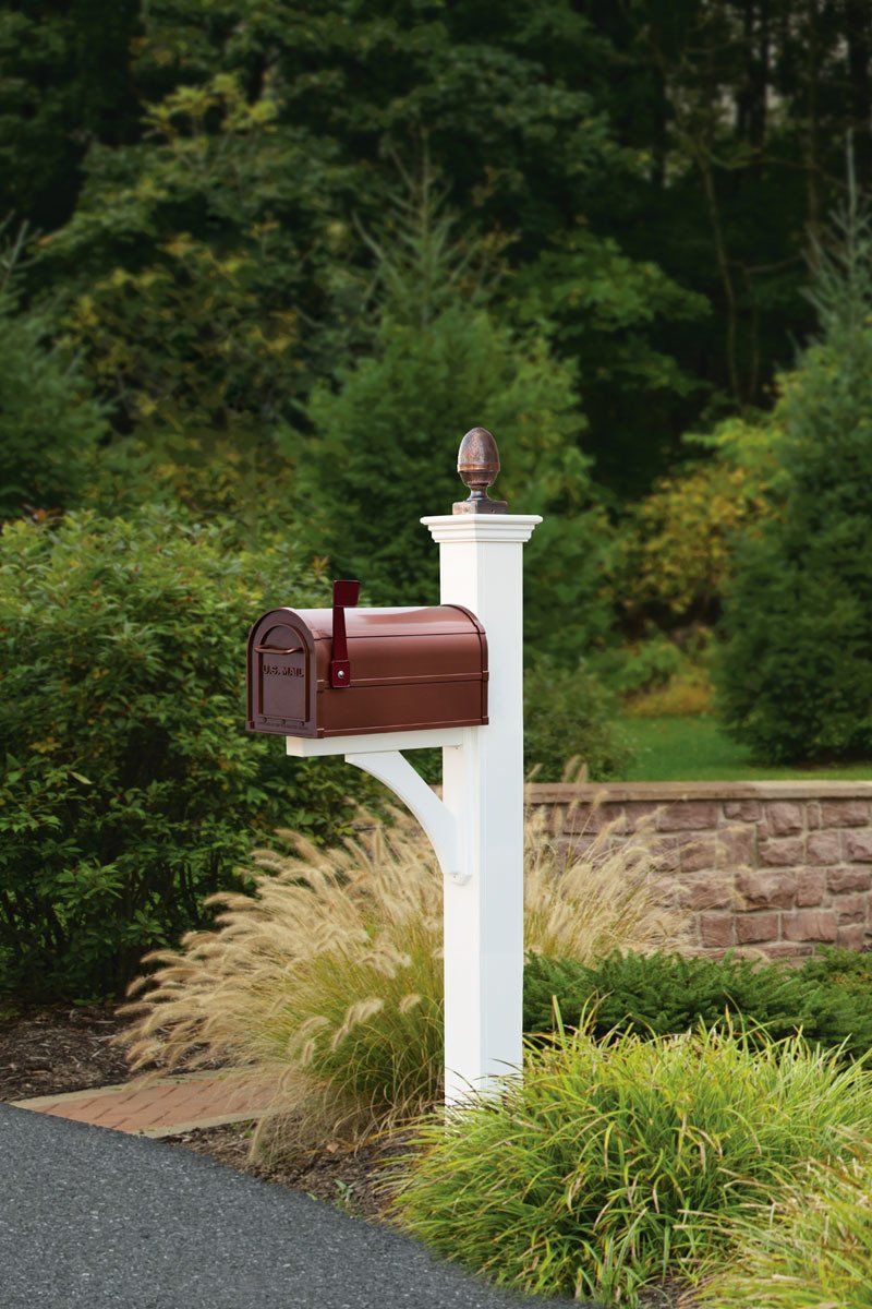 A brown mailbox is sitting on top of a white post next to a driveway.
