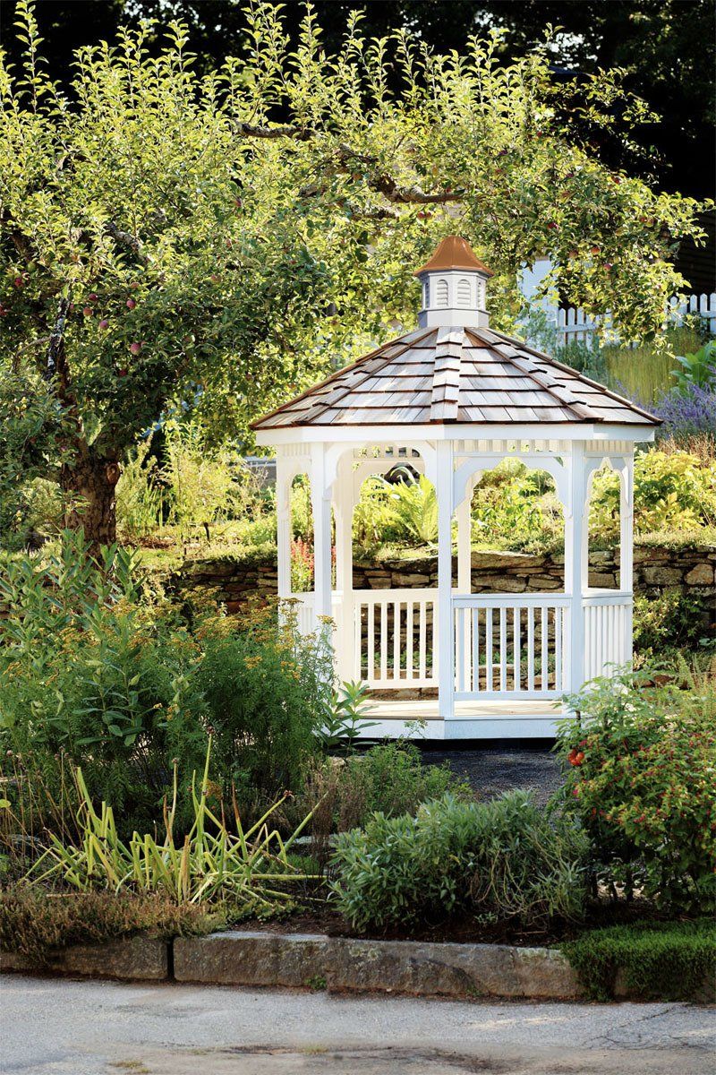 A white gazebo is surrounded by trees and bushes in a garden.